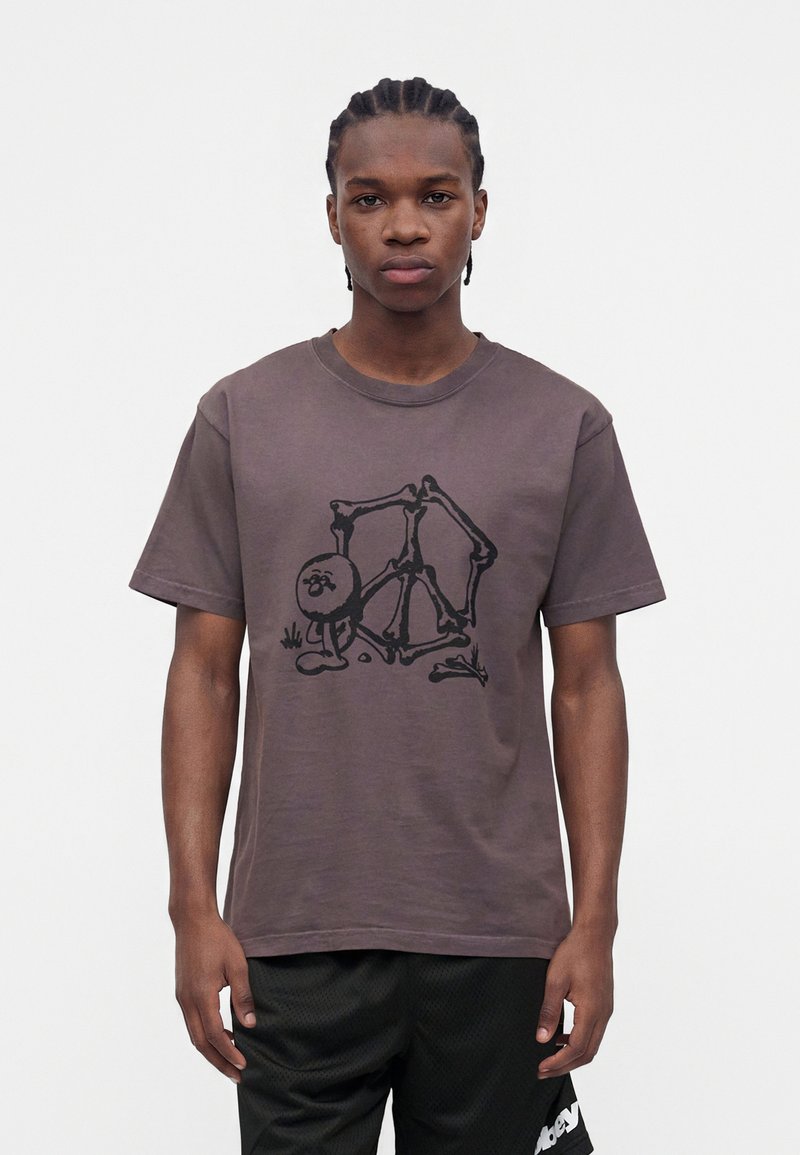 Νέος άνδρας με πλεξούδες, που φοράει καφέ t-shirt με μαύρο αφηρημένο σχεδιασμό σκελετού και μαύρα αθλητικά σορτς.