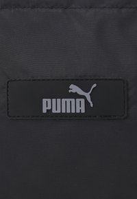 Czarny materiał nylonowy o teksturowej powierzchni, z prostokątną czarną etykietą z gumy, na której znajduje się napis "PUMA" oraz szare logo skaczącego kota.