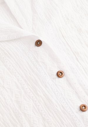 Material alb texturat cu un model în formă de diamant, cu nasturi maro detaliind partea din față. Material moale și ușor.