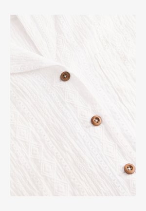 Tissu blanc texturé avec un motif en losanges, orné de boutons marron sur le devant. Matière douce et légère.