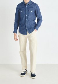 Dark denim shirt with two chest pockets, light beige pants, and navy sneakers with white soles, set against a plain background.