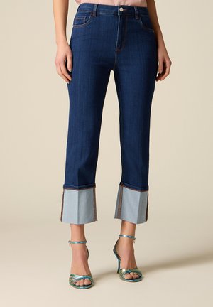 Donna che indossa jeans corti blu scuro con ampi risvolti piegati di colore azzurro chiaro e sandali metallizzati con cinturini e tacco alto.