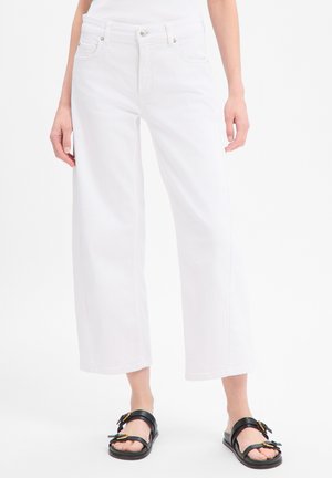 Vrouw draagt witte cropped wijde jeans en zwarte sandalen met gesp, staand tegen een effen achtergrond.