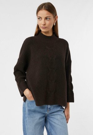 Brązowy sweter dzianinowy z wysokim kołnierzem, teksturowanym wzorem prążkowym i szerokimi rękawami, w połączeniu z jasnoniebieskimi dżinsami.