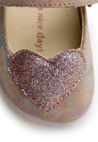 Zapato de gamuza color canela con un acento en forma de corazón brillante en tonos multicolores. Presenta una puntera redondeada y una correa asegurada. Impresión de "¡que tengas un buen día!" en el interior.