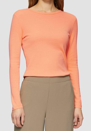 Kvinde iført en langærmet ferskenfarvet top og højtaljede beige bukser, stående mod en ensfarvet lys baggrund.