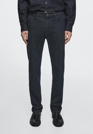 Homme portant un jean en denim foncé, des mocassins en cuir noir et une chemise boutonnée foncée, debout devant un fond uni.