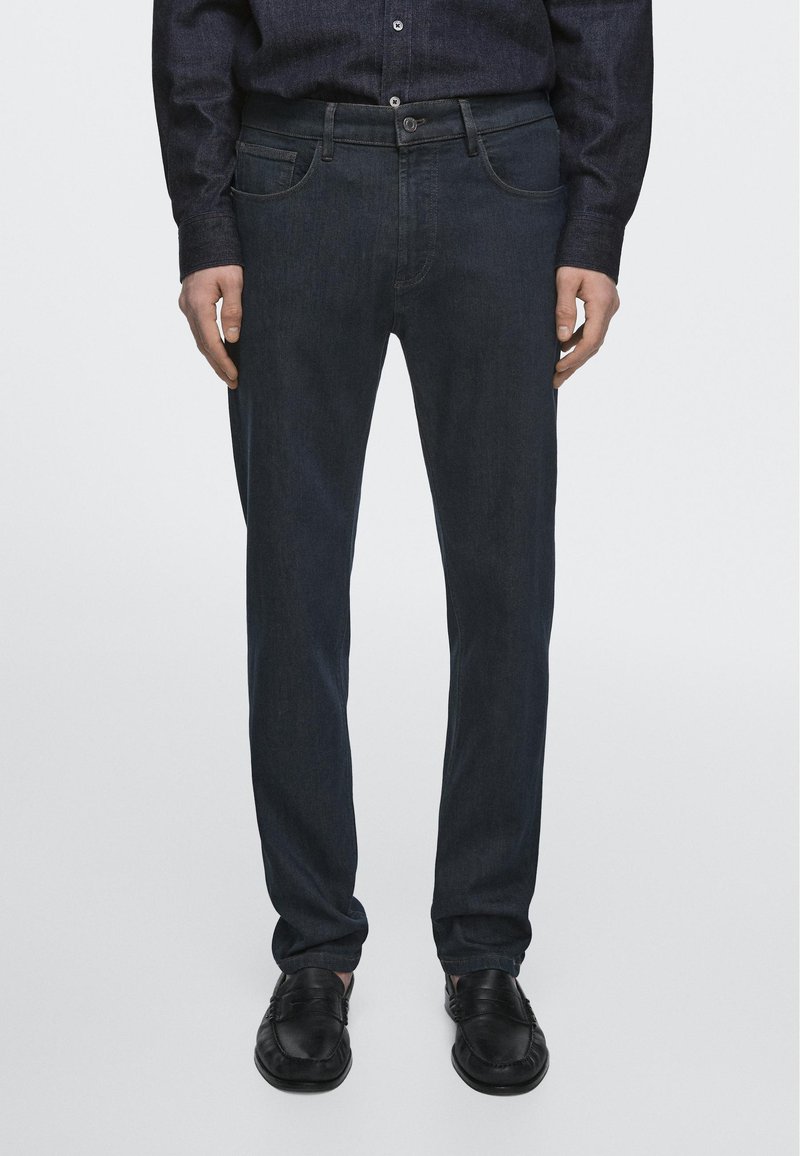 Man die een donkere spijkerbroek, zwarte leren loafers en een donker blouse met knopen draagt, staande voor een effen achtergrond.