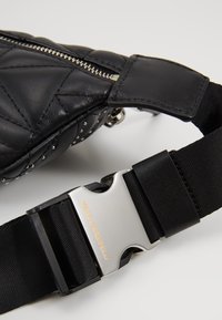 Mustekattu musta nahkakassi, jossa on hopeiset niittiyksityiskohdat ja musta hihna, johon on kaiverrettu hopeinen solki, jossa lukee "Karl Lagerfeld."