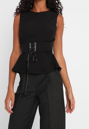 Mujer con top negro sin mangas tipo peplum con cinturón de cintura ajustable con cordones y pantalones anchos de color gris oscuro, sobre fondo liso.