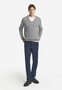 Grauer gestrickter Pullover mit V-Ausschnitt, über einem weißen Hemd getragen. Kombiniert mit dunkelblauen geraden Jeans und schwarzen Sneakers.