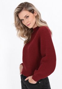 Burgundowy sweter z prążkowanej dzianiny o wysokim kołnierzu i bufiastych długich rękawach. Noszony z ciemnymi dżinsami, prezentując teksturalny, przytulny design.