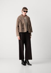 Persoon in een oversized bruine wollen jas, beige trui, zwarte wijde broek, zwarte haklaarzen en zwarte zonnebril tegen een witte achtergrond.