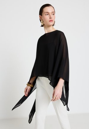 Donna che indossa una maglia nera trasparente asimmetrica, pantaloni bianchi, orecchini d'oro e braccialetti su uno sfondo chiaro e uniforme.