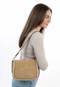 Beżowa torebka crossbody z ekoskórki z czarnymi przeszyciami, regulowanym paskiem, gładką teksturą i zapięciem na klapkę, prezentowana na modelce.