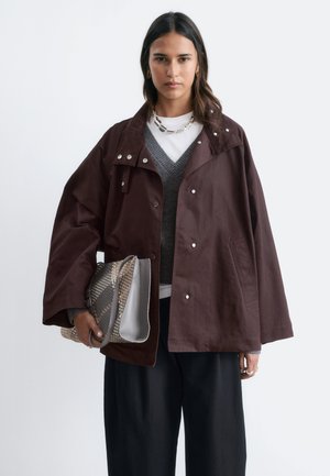 Femme aux longs cheveux foncés porte une veste marron oversize sur un pull gris et une chemise blanche, tenant une grande pochette texturée.