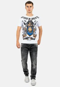 Maglietta grafica bianca con una stampa audace e accenti neri e colorati; abbinata a jeans grigi scuri e sneakers bianche.