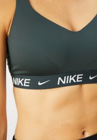Sujetador deportivo gris oscuro con un diseño de escote en forma de V, que incluye un logo blanco de "NIKE" y una banda suave y elástica para soporte. Tejido liso y elástico.