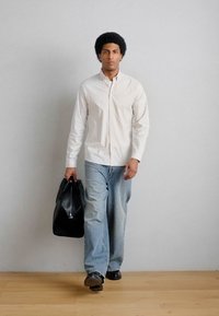 Homme marchant à l'intérieur portant une chemise blanche rayée et un jean ample, tenant un grand sac en cuir noir dans sa main gauche.
