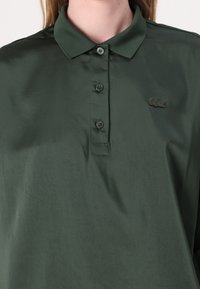 Mørk grønn polo skjorte laget av glatt stoff, med krage, fire knappestolpe og brodert logo på venstre bryst.