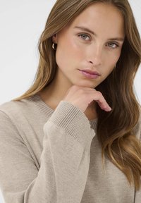 Pull en maille beige avec des poignets et un col en côtes. Texture lisse, design semi-ajusté. Modèle reposant sa main sur le menton, avec de longs cheveux visibles.