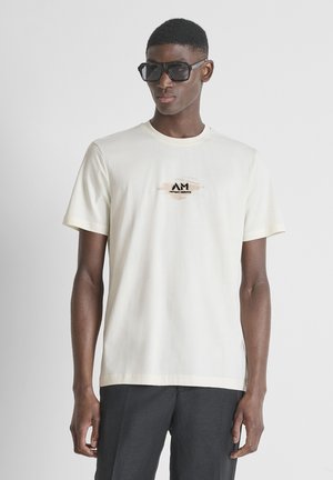 Joven con gafas de sol negras, camiseta de manga corta color crema con un pequeño logo y pantalones oscuros, posando frente a un fondo claro y sencillo.