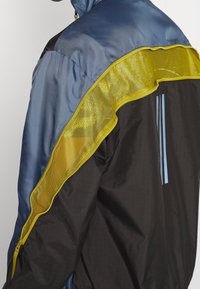Sportjacke mit einem oberen Teil aus blauer Satin, schwarzem Körper und gelben Mesh-Akzenten. Bemerkenswerte Details sind seitliche Reißverschlüsse und vertikale Streifen.