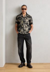 Camicia nera a maniche corte con motivo e dettagli gialli, abbinata a jeans in denim scuro e stivali alla caviglia marroni. Modello che indossa occhiali da sole.