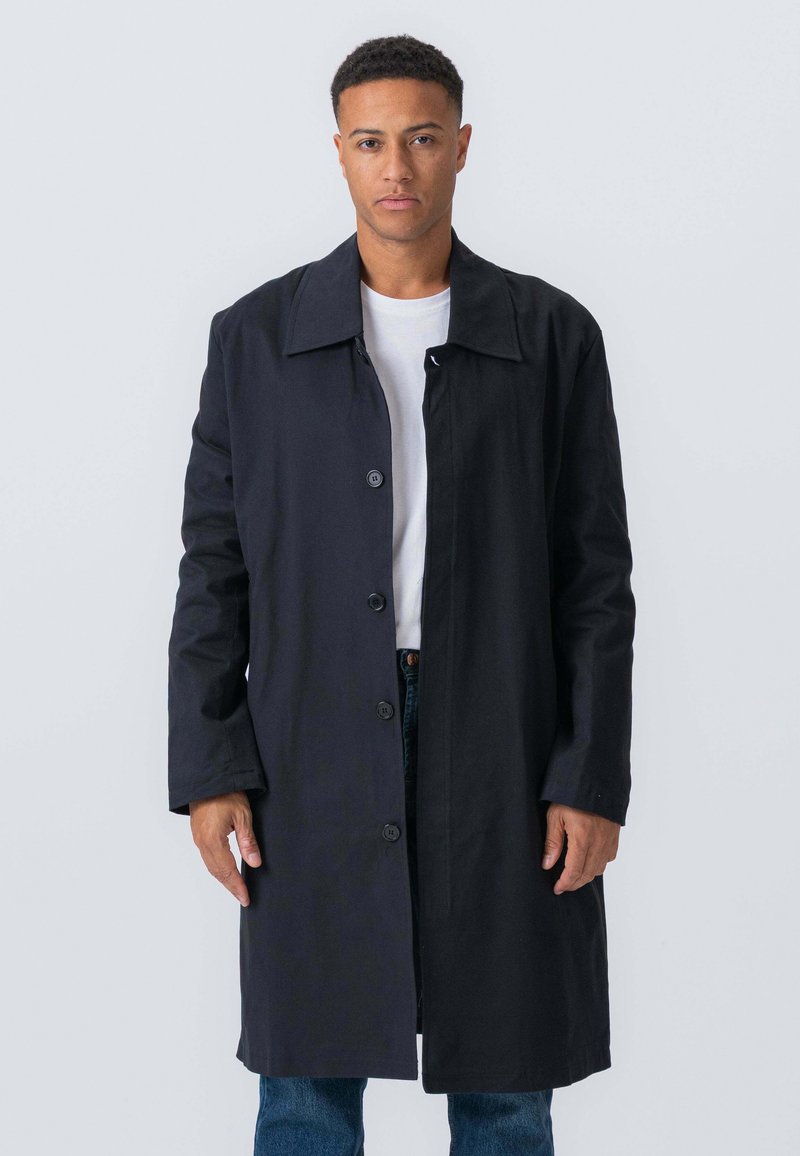 Mand står oprejst iført en lang sort knapjakke over en hvid skjorte og blå jeans, vender mod fronten med en neutral ansigtsudtryk.