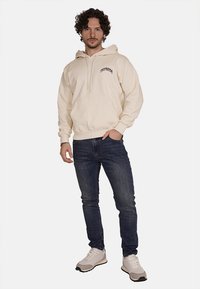 Felpa color crema con tasca frontale, cappuccio con cordoncino e logo ricamato; abbinata a jeans scuri e sneakers bianche.