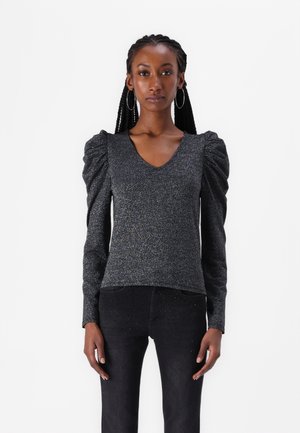 Zwarte, glinsterende top met lange mouwen, een V-hals en pofmouwen. Gekleed met zwarte jeans met subtiele versieringen.