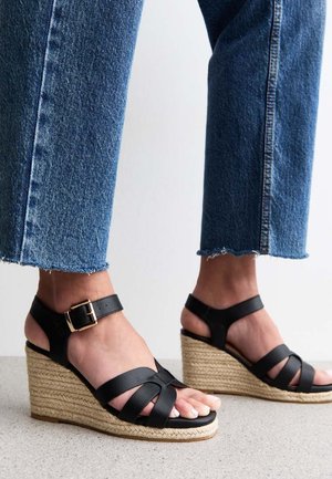 Sandalias de cuña negras con correas entrecruzadas y hebilla en el tobillo, con una suela trenzada de yute. Llevadas con pantalones vaqueros azules recortados.