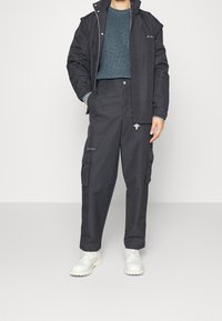 Homme portant un pantalon cargo noir, une veste noire, un pull en tricot gris et des bottes blanches se tenant contre un fond uni.