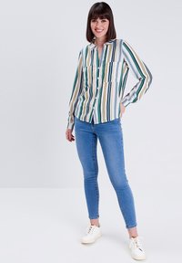 Blouse rayée avec des lignes vertes, jaunes et blanches, manches longues, fermeture à boutons et poches poitrine doubles, associée à un jean skinny bleu et des baskets blanches.