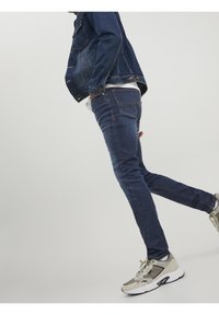 Jeans de mezclilla oscura con ajuste slim, combinados con una chaqueta de mezclilla y zapatillas gris claro. Incluye detalles de costura y un diseño clásico.