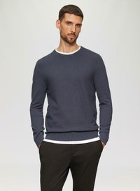 Marineblauer Strickpullover mit Rundhalsausschnitt, strukturiertem Stoff und gerippten Bündchen. Über einem weißen Shirt getragen, das am Ausschnitt hervorblitzt.