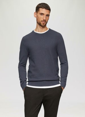 Marine gebreide trui met een ronde hals, textuurstof en geribbelde mouwen. Gelaagd over een wit shirt dat iets zichtbaar is bij de halslijn.