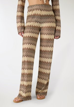 Persona che indossa pantaloni a maglia con motivo a zigzag beige e marrone e top a maniche lunghe abbinato con infradito, in piedi davanti a uno sfondo bianco.