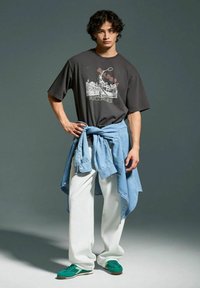 Maglietta grafica nera, camicia di denim azzurro chiaro annodata in vita, pantaloni bianchi larghi e sneakers verdi indossati da un modello su uno sfondo grigio.