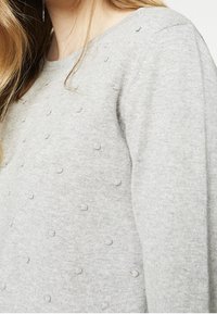 Nainen, joka on pukeutunut vaaleanharmaaseen neulepaitaan, jossa on pieniä kohopisteitä, näkyy olkapäästä vyötärön puoliväliin saakka.