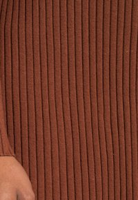 Tissu tricoté marron à côtes avec des rayures verticales, mettant en avant une texture douce et un espacement uniforme dans le design. Une main partiellement visible.