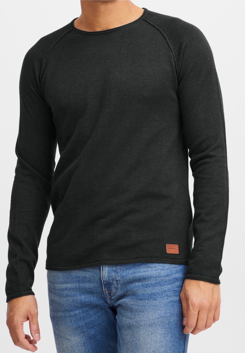 Chemise noire à manches longues avec col rond, texture côtelée, et une étiquette logo en cuir marron à l'ourlet, associée à un jean denim bleu.