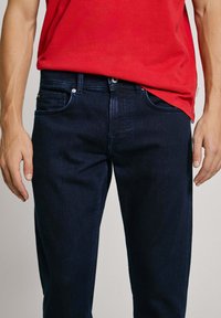 Dark blue denim jeans with a straight cut, five pockets, and metal button closure. The fabric has a smooth texture and minimal fading.
