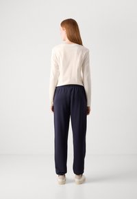 Žena otočená zády, oblečená v krémovém tričku s dlouhým rukávem, tmavě modrých kalhotách s elastickým pasem a béžových teniskách na jednobarevném světlém pozadí.