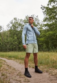 Lichtblauw langemouwen top met een rits aan de voorkant, gecombineerd met lichtgroene shorts en zwarte sport schoenen. Natuurlijke omgeving met gras en bomen.