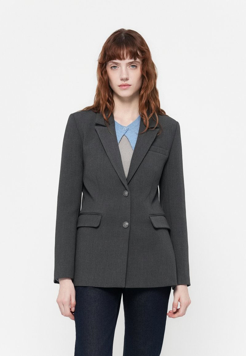 Blazer grigio su misura con revers a notch, due tasche frontali, chiusura con un bottone e trama liscia. Indossato sopra una camicia blu con colletto.
