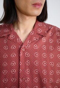 Camicia rossa a maniche corte con motivo di cuori bianchi e pois, colletto a punta e chiusura con bottone al collo.