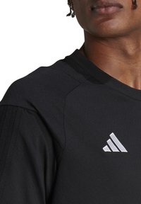 Närbild av en person som bär en svart Adidas sporttröja med den vita Adidas-logotypen på bröstet och texturerade randar på ärmen.