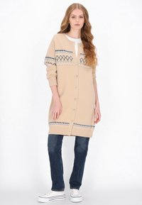 Beige neulottu neuletakki-mekko, jossa on napitus edessä. Rinnassa ja hihansuiden resoreissa sinivalkoinen geometrinen kuvio.