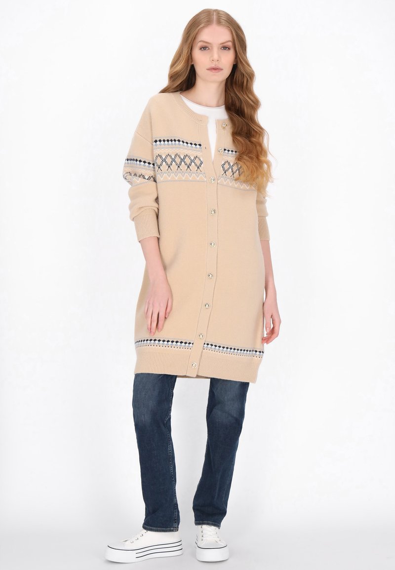 Beige neulottu neuletakki-mekko, jossa on napitus edessä. Rinnassa ja hihansuiden resoreissa sinivalkoinen geometrinen kuvio.